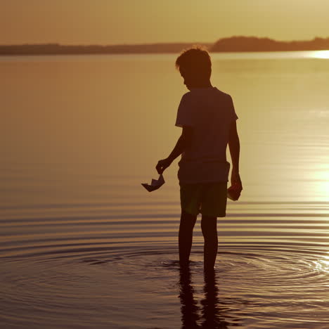 boy with red and green paper ships is walking in the river. origami paper boats in kid's hands. beautiful waves ripple background. travel and nature concept. video at sunset.