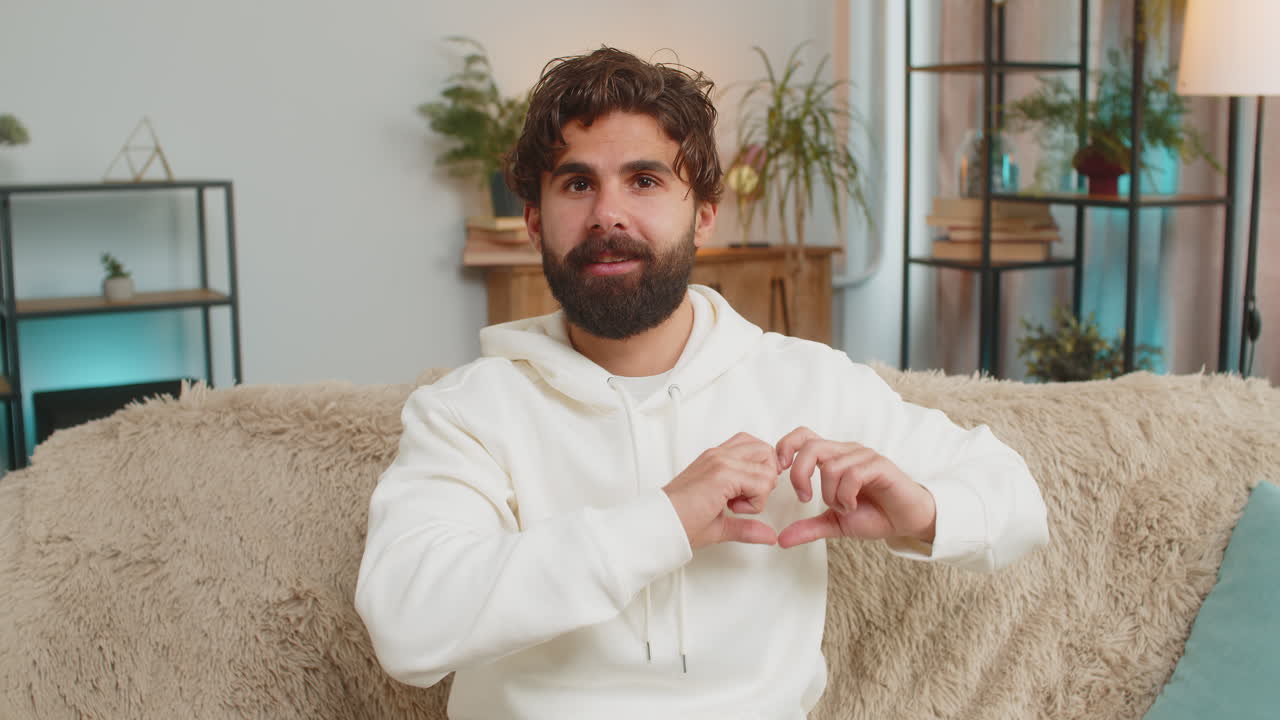 행복한 인도인 청년은 집에서 거실 소파에서 사랑의 상징을 만니다. 카메라에 심장 기호를 보여줍니다.