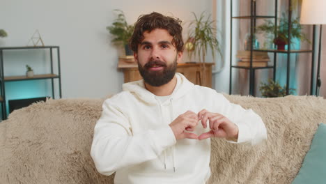 joven indio feliz en casa sofá de la sala de estar hace símbolo de amor mostrando el corazón señal a la cámara