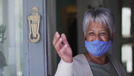 Senior-mixed-race-woman-wearing-face-mask-opening-front-door-waving-greeting