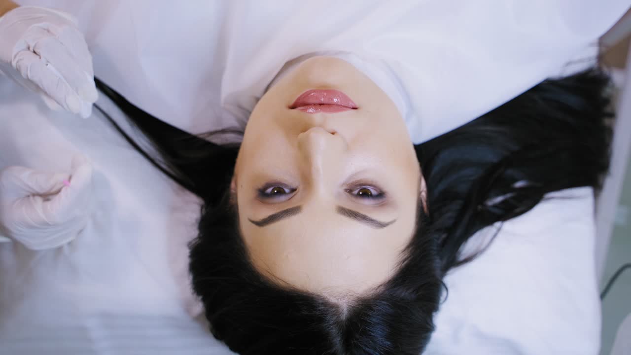 Specialist dries with a sponge lips to a woman after the permanent make-up. Top view of a beautiful female's face in a beauty salon. Slow motion