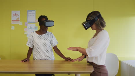 two diverse female coworkers standing at desk, testing vr googles