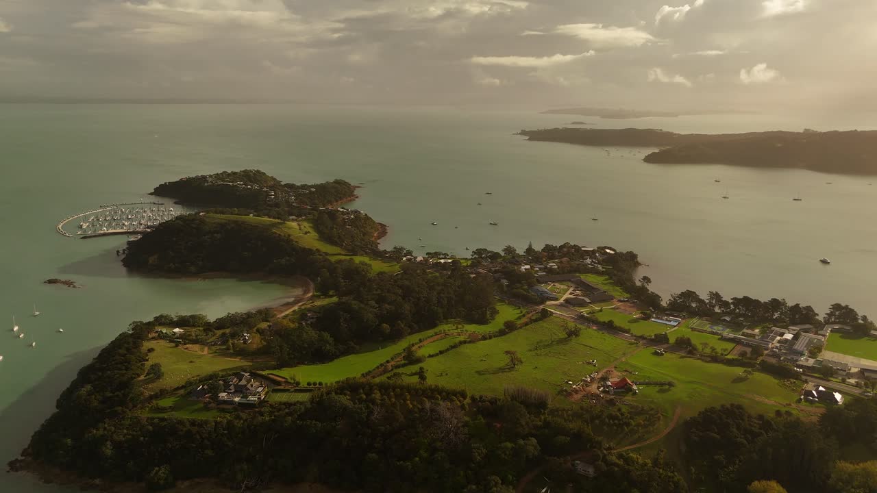 Green hills with coastline and port harbor with boats during cloudy sunset. Aerial wide shot. New Zealand, Haiheke Island, Surfdale Area. Shelly Beach.