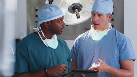 Two-Male-Surgeons-Wearing-Scrubs-Looking-At-Digital-Tablet-In-Hospital-Operating-Theater