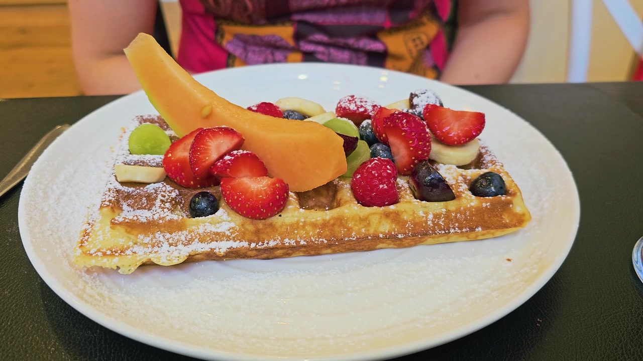 Waffle dolce alla frutta su un piatto bianco con fragole, melone, banana, mirtilli, uva, lamponi e zucchero a velo, ripreso da vicino come gustoso cibo da caffè per brunch o dessert.