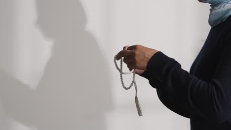 muslim woman wearing hijab praying holding prayer beads casting shadow on wall behind