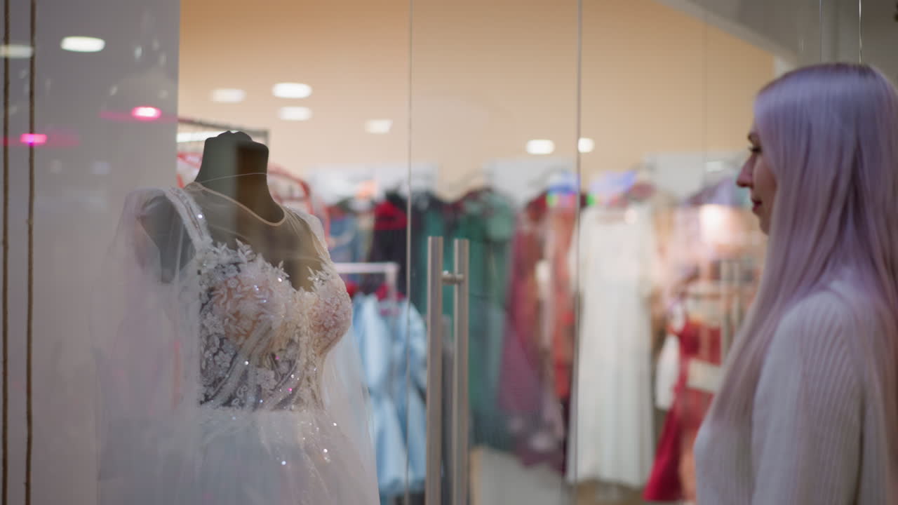 beautiful wedding gown behind glass display in mall boutique as lady stands admiring intricate lace details and sparkling embellishments, soft overhead lighting reflecting on glass, mannequins