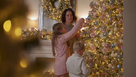 little boy and girl decorate christmas tree with balls and ornaments, mother lifting them up