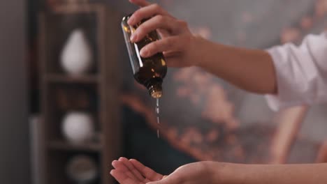 the cosmetologist or massage therapist in the beauty salon's room holds a dark jar of beauty treatment fluid and pours it onto their hands in the air