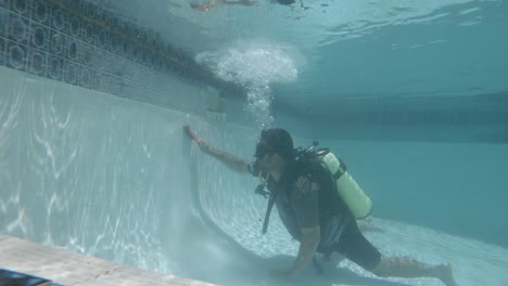 a professional pool cleaner in scuba equipment scrubs dirt and algae underwater from the side of a pool with a brush