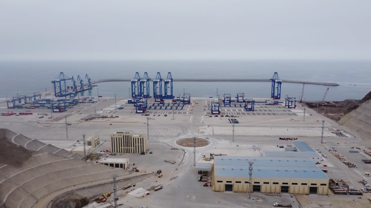 Aerial shot of a new Megaport under construction in the city of Chancay, Peru. Drone slowly orbits around. Many blue cranes can be seen on the edge of the harbor and behing the ocean horizon.