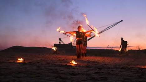 a group of men and woman fire show at night on the sand against the background of fire and tower cranes