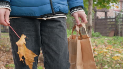 lower angle view of kid standing holding brown paper bag with autumn leaf in right hand, turning it clockwise and counterclockwise, with blurred background of bar fence and fallen leaves