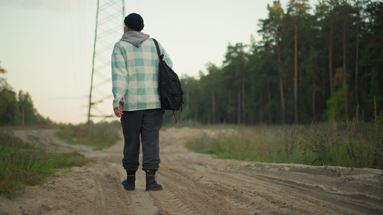 Escursionista caucasico che si allontana lungo la strada, zaino in spalla, passo fermo verso l'orizzonte della foresta, cielo crepuscolare che si riscalda, stato d'animo risoluto, viaggiatore solitario che prosegue, viaggio nella natura selvaggia, partenza piena di speranza
