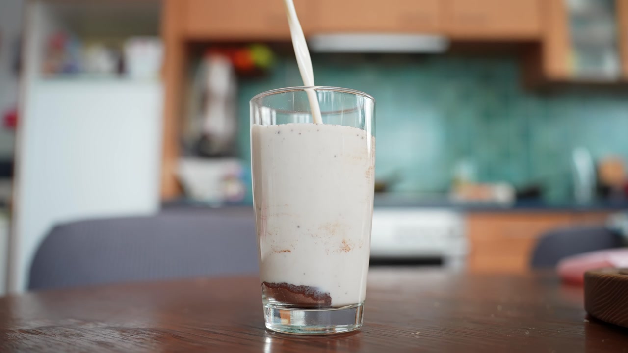 Milk pours into a tall glass with cocoa powder at the bottom, starting homemade chocolate milk on a wooden table with a blurred kitchen background.