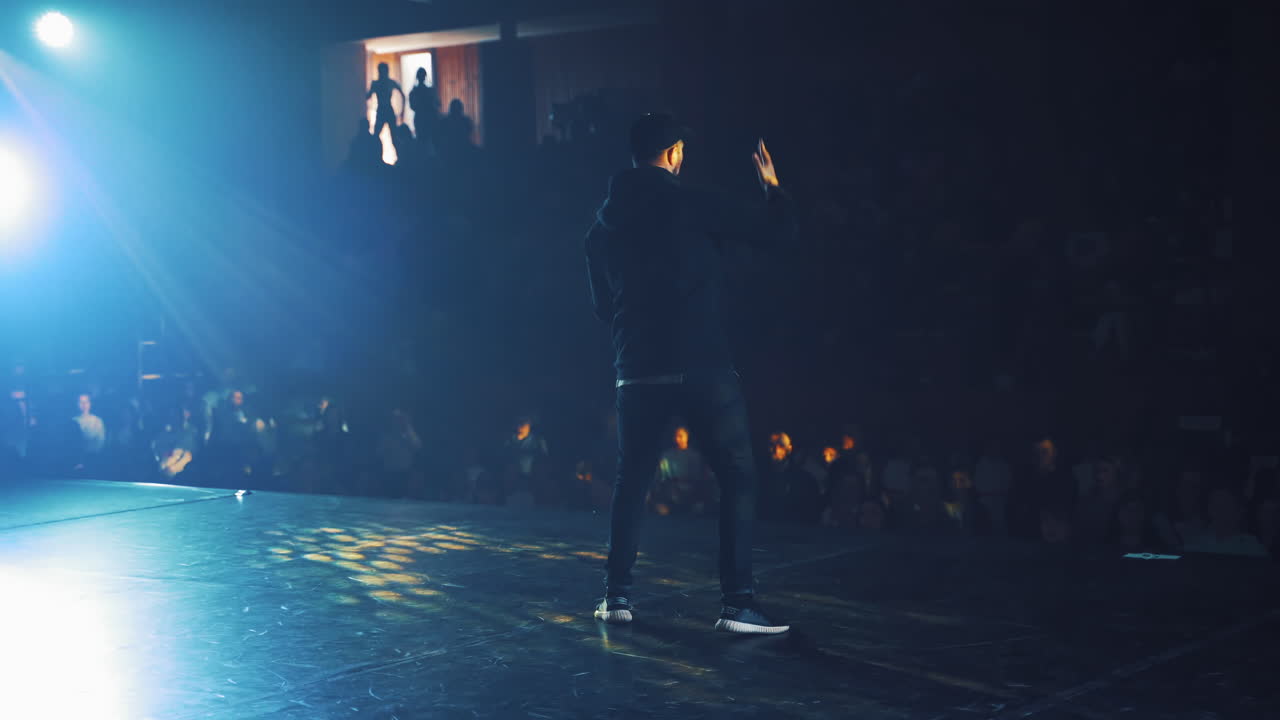 Hip-hop dancer in black cap and casual clothes dances on stage in the club. Modern dance class on scene for young audience in the studio indoors. Dancing concert.