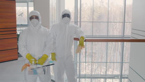 two cleaning men wearing personal protective equipment looking at camera inside an office building while holding cleaning products and rags