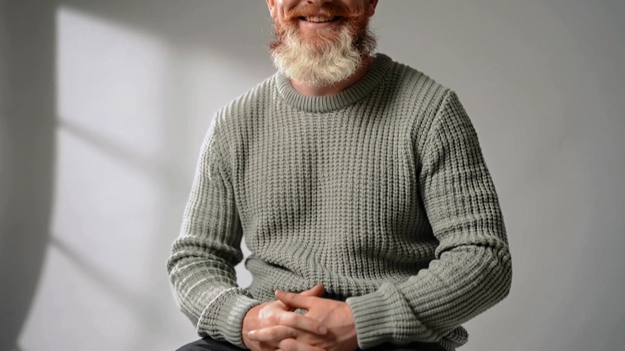 La telecamera si sposta verso il basso rivelando di più, uomo con la barba che stringe un bicchiere seduto su uno sgabello, maglione verde salvia, ombra a sinistra