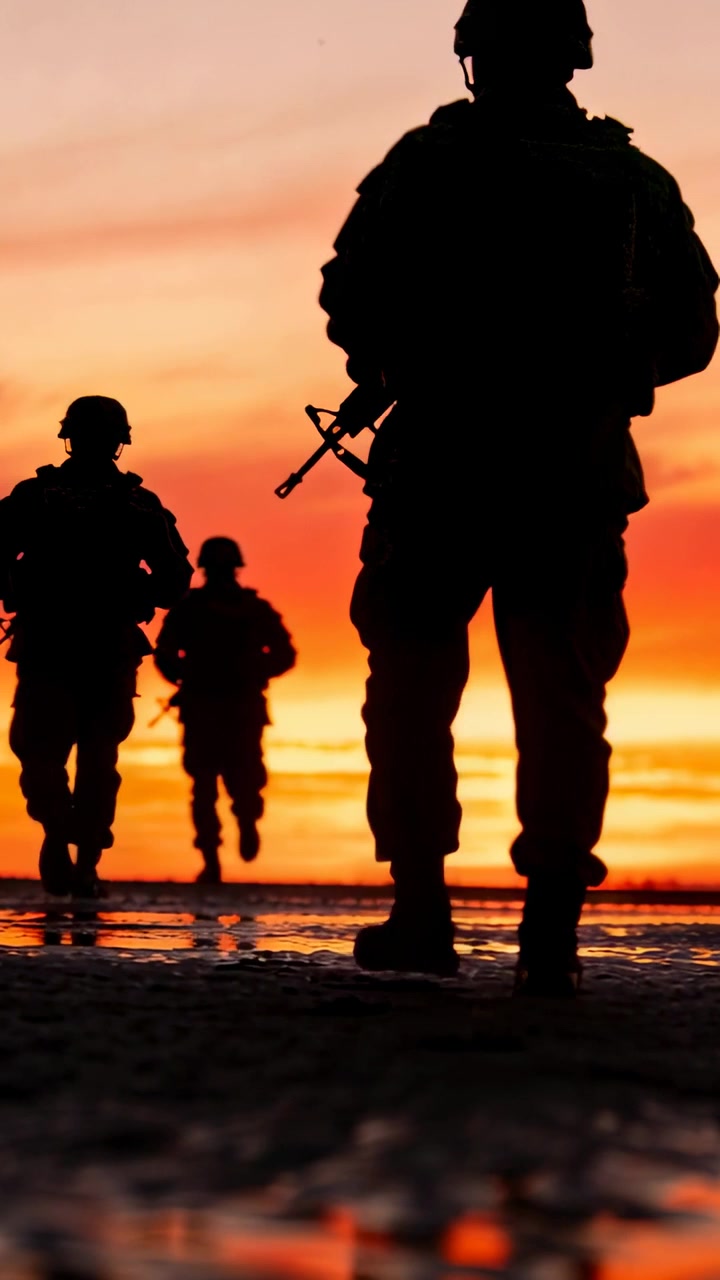 Vertical video: Walking front soldier advancing toward camera on wet sand with helmet, vest, rifle