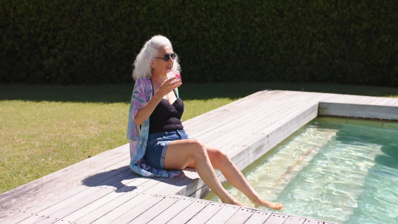 Mature woman, swimsuit and sunglasses, relaxing on pool deck, raising pink drink, smiling at camera