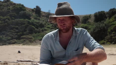 A-man-in-a-blue-shirt-and-a-hat-reading-a-bible-on-the-beach