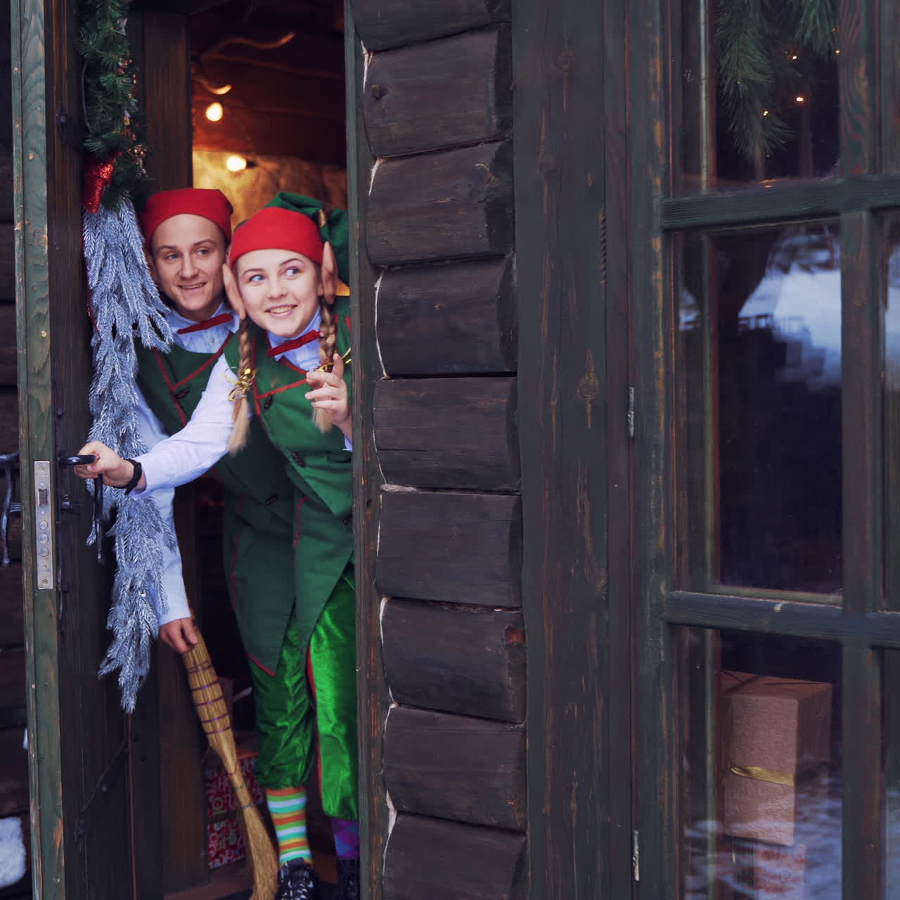Wooden door opens and two elves looking in surprise. Curious fairy characters in green costumes peeking out inside the house. Christmas time.