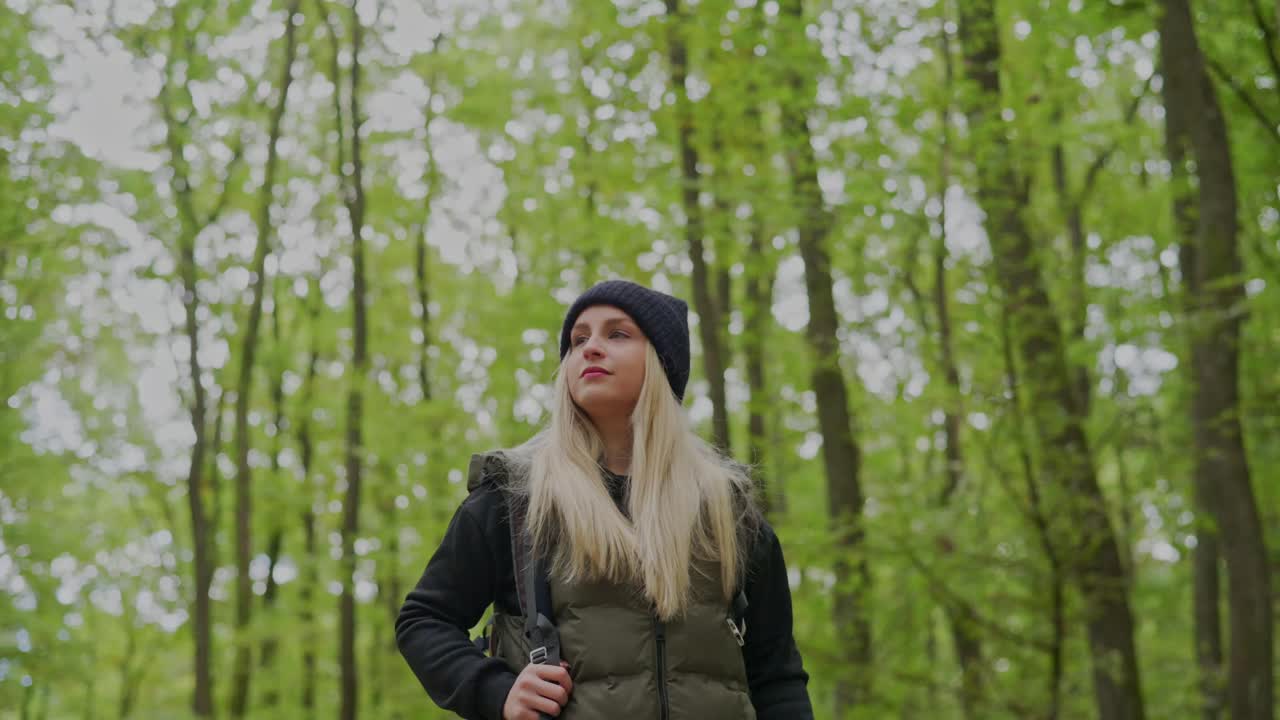 A young woman wearing a beanie and warm jacket stands in a forest surrounded by tall green trees. She carries a backpack and looks thoughtfully into the distance, enjoying a peaceful hike in nature