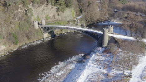 Aerial-view-of-Craigellachie-bridge-surrounded-by-snow-on-a-sunny-winters-day-with-a-lone-person-standing-on-the-bridge,-Moray,-Scotland---receding-and-rising-shot