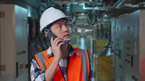 close up of asian male engineer with safety helmet talking on smartphone while standing in engine control room, work of electrical generators