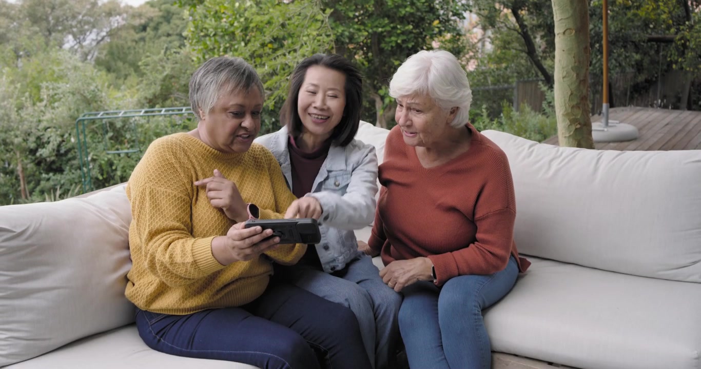 Diverse women including senior on deck sofa, left woman holding device, others tapping and viewing