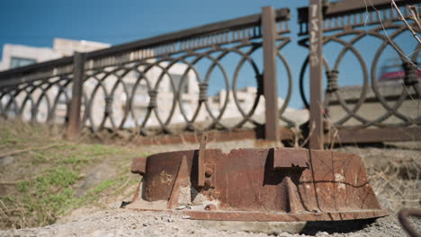 close view of a rusty metal object lying on the ground with an ornate urban fence in the background, with cars passing on the road