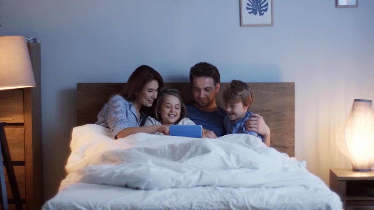 Caucasian parents sitting on the bed with their little cute son and daughter, watching something on the tablet and talking at night