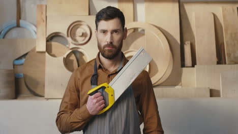 portrait of caucasian bearded man in helmet and holding saw and looking at camera in carpentry workshop