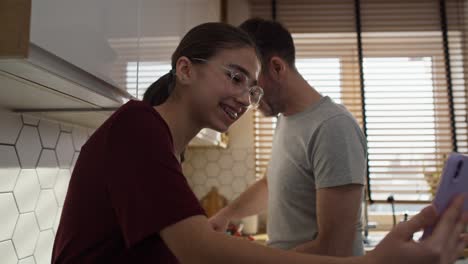 caucasian father with teenage daughter looking at mobile phone in kitchen while cooking