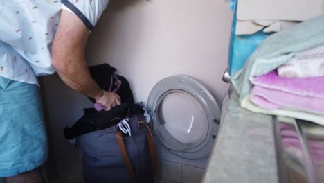 wide shot of man brining a clothes basket into the laundry and putting a load of clothes on in a front load washing machine, sorting clothes into the machine by colour