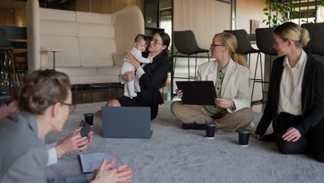 Close-up-five-confident-business-women-in-business-suits-sit-on-the-carpet-in-a-modern-office-and-communicate-while-one-of-them-holds-a-small-child-in-her-arms