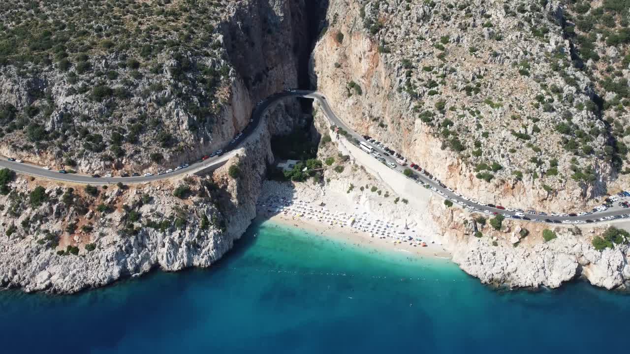 Aerial view of coastal road curving over tunnel beside white sand beach and turquoise sea near Kaputas Beach in Antalya Province, Turkey, surrounded by steep rocky cliffs and clear water