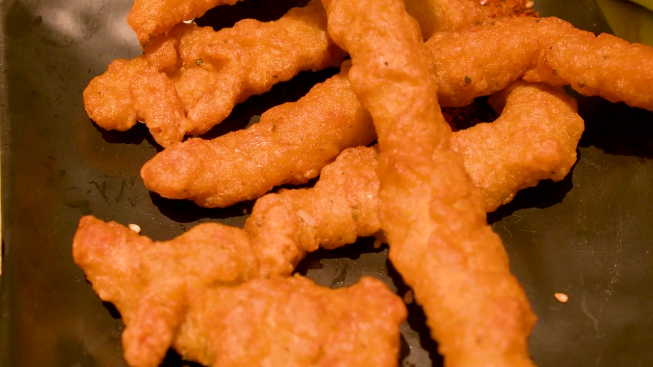 Primo piano di bacchette che sollevano pollo tempura dorato da un piatto nero con illuminazione calda.