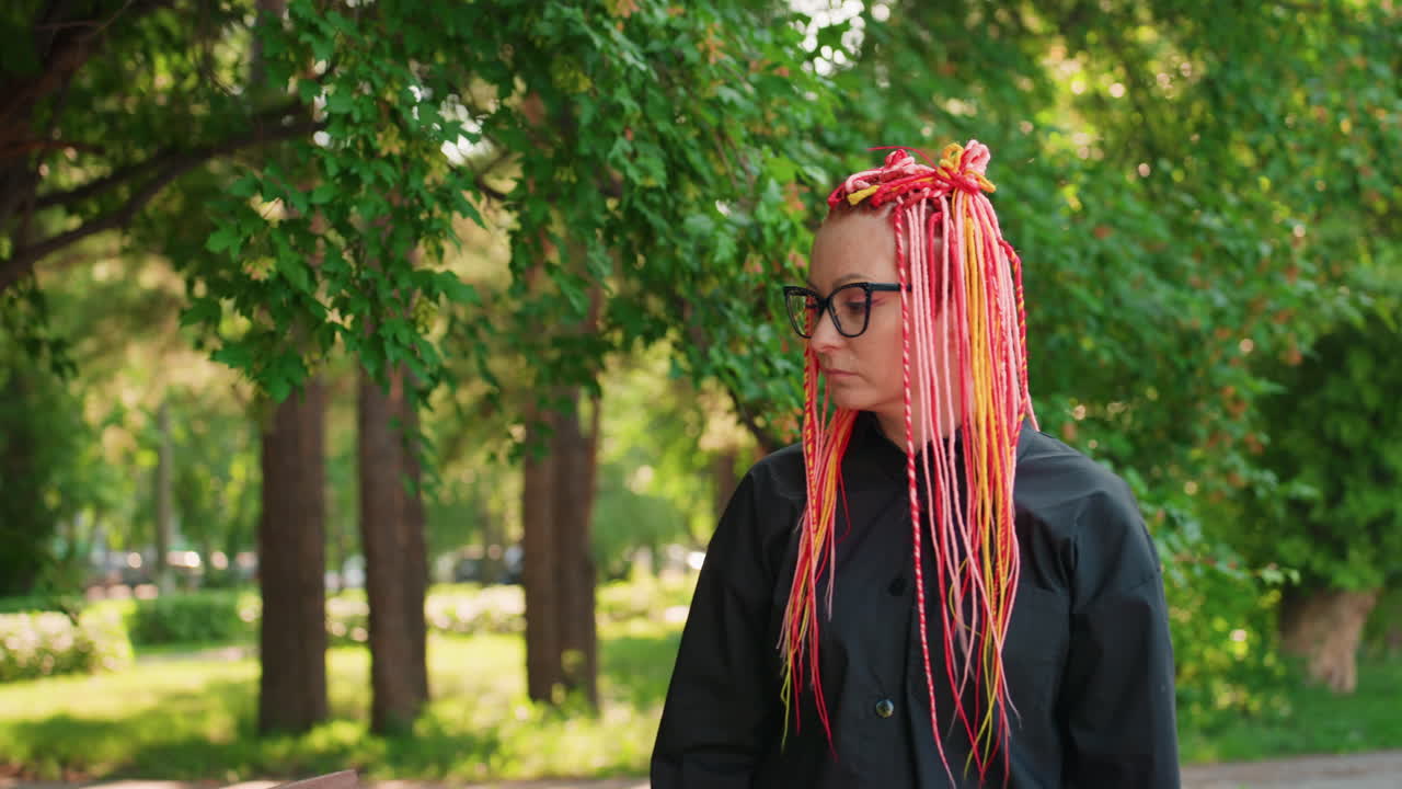 주황색과 분홍색 땋은 머리를 한 백인 기업가가 공원 벤치에 앉아 팔을 판자에 걸치고, 흔들리지 않는 시선과 차분한 자세로 잎이 무성한 거리와 부드러운 햇빛 아래에서 다음 크리에이티브 프로젝트를 기획하고 있어요.