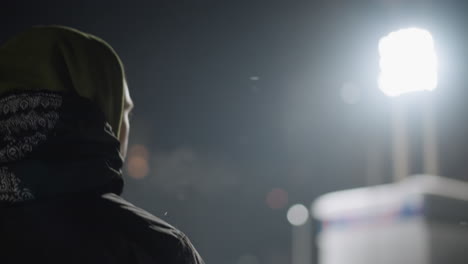 back view of man wearing hoodie and patterned jacket facing bright stadium lights on cold winter night, soft snow falling in dim surroundings with distant light flares and urban winter atmosphere