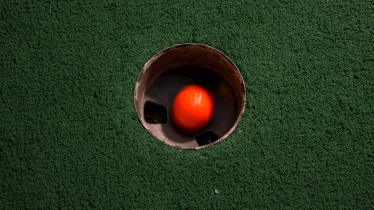 A close up birds eye view of an orange mini golf ball falls into the golf hole and bounces in the pocket on a course