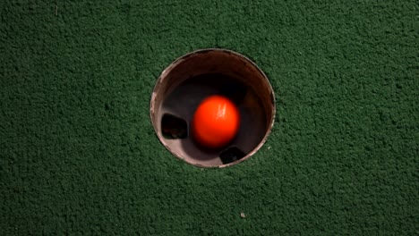 una vista de pájaro de cerca de una mini pelota de golf naranja cae en el hoyo de golf y rebota en el bolsillo en un campo