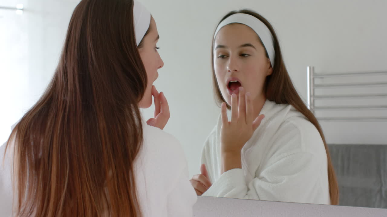 Applying skincare product, woman in bathrobe looking in mirror at home spa