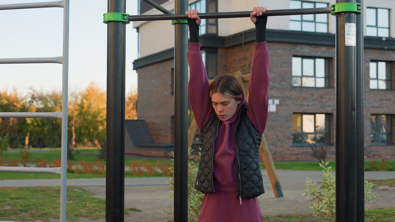 Workout junkie grips horizontal bar for balance in urban outdoor gym, showcasing forearm strength, focus, and control on rubber flooring with autumn foliage and city building backdrop