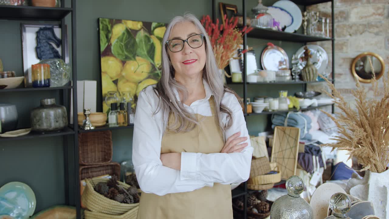 Portrait of adorable smiling happy mature woman with grey hair in glasses which poses on camera with crossed arms in beautiful presents shop