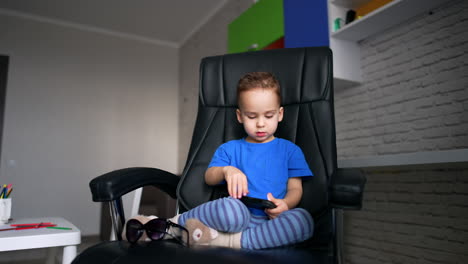 charming caucasian boy sits in the office chair. sweet baby is focused on the smartphone he's holding in hands.