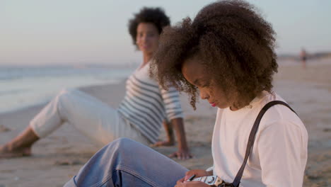 Nahaufnahme-Eines-Lächelnden-Mädchens,-Das-In-Die-Kamera-Schaut,-Während-Es-Zeit-Am-Strand-Mit-Seiner-Mutter-Verbringt,-Die-Im-Verschwommenen-Hintergrund-Sitzt