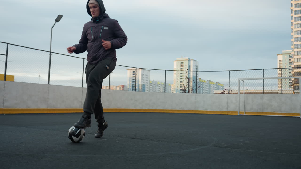 atleta entrenando hábilmente con el fútbol en la arena deportiva, tocando la pelota hacia adelante con precisión y control, el fondo incluye cercas en forma de cadena, edificios residenciales y cielo abierto