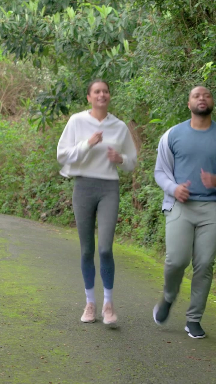 Video verticale: Due persone che fanno jogging su un sentiero in collina, con una donna che rallenta, si piega e si allunga per recuperare, controllando l'orologio.