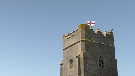 Englischer-Kirchturm-Mit-Englischer-Flagge-Auf-Blauem-Himmelshintergrund,-Zeitlupe-Mit-Wind